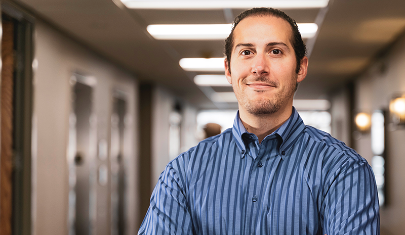 A man in a blue striped shirt standing confidently in a well-lit hallway with a neutral expression.