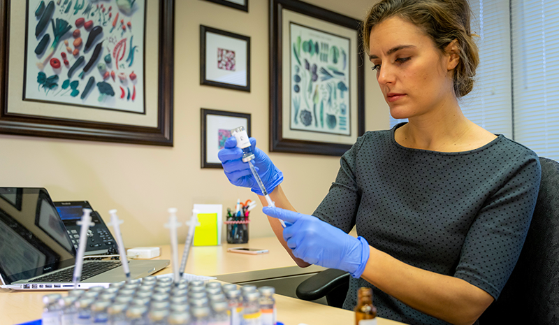 A woman wearing gloves and a professional outfit preparing a syringe in an office setting, surrounded by vials and framed artwork on the wall.