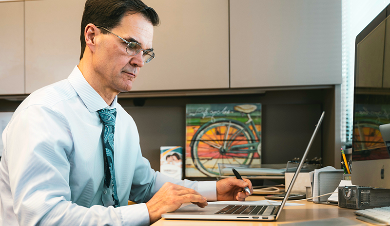 A man in professional attire working at a desk with a laptop and a pen, with a colorful bicycle painting in the background.